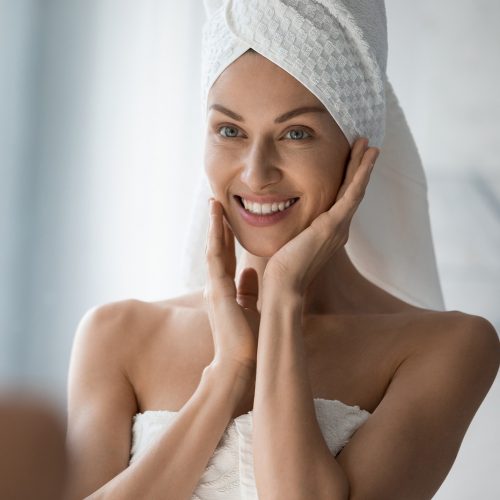 After shower body and head wrapped in towel 35s woman looks in mirror touches moisturized soft healthy face skin feels satisfied enjoy spa cosmetics treatment procedure, morning care hygiene concept